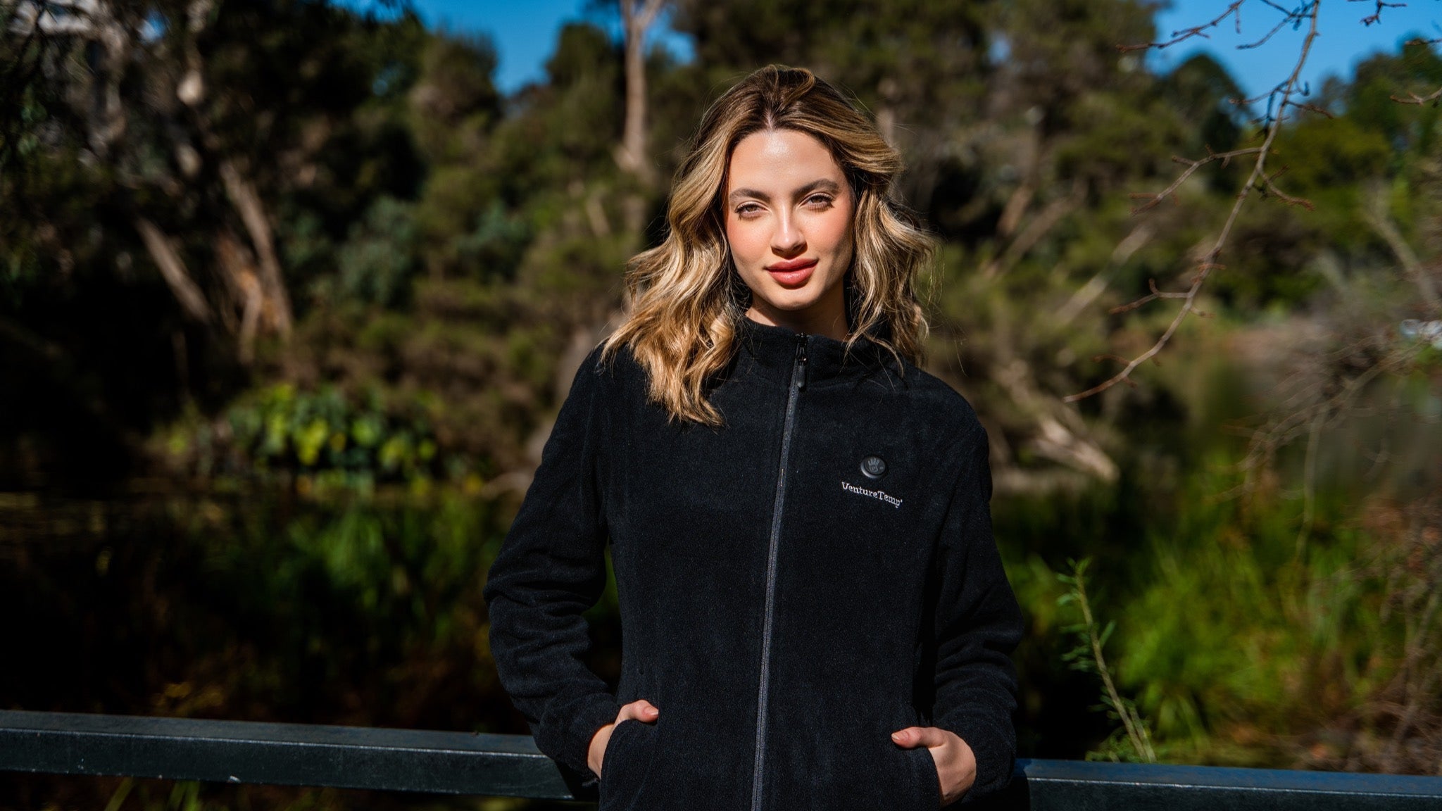Woman wearing a black heated jacket with a logo, standing outdoors with greenery in the background